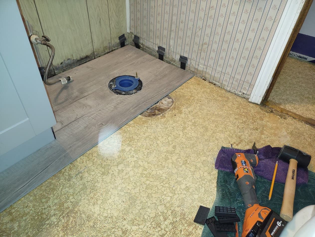 Gray wood-look vinyl plank flooring laid up to the toilet flange over the old linoleum, with perimeter spacers and install tools set out