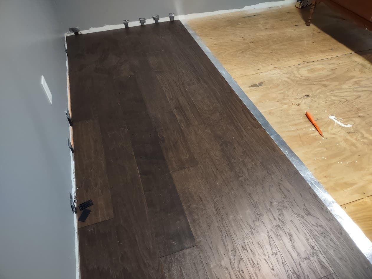 Dark hardwood planks going down over the prepped subfloor, with perimeter spacers holding the expansion gap