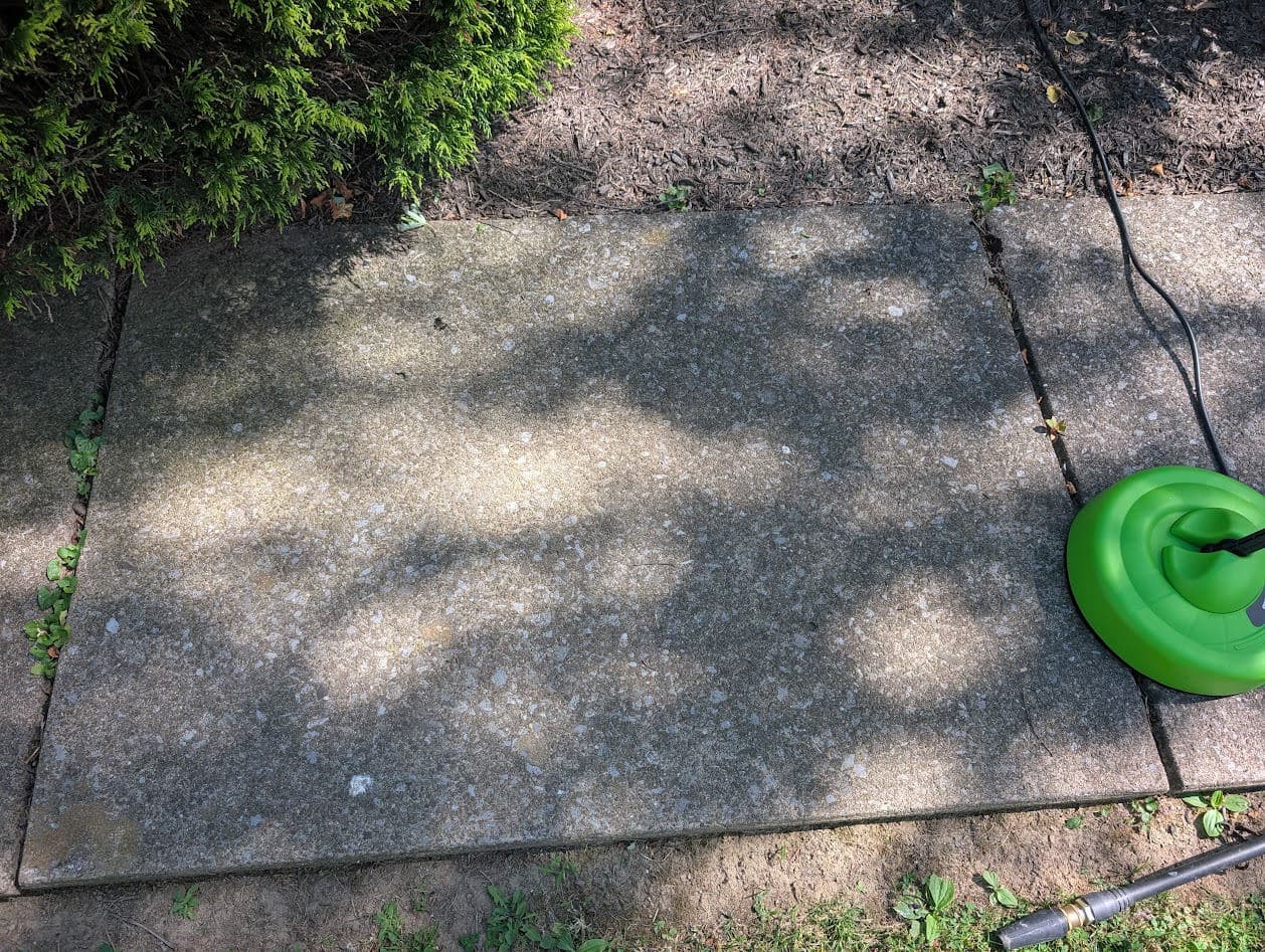 The exposed-aggregate concrete sidewalk before cleaning, darkened with algae, with a pressure-washer surface-cleaner attachment resting at the edge